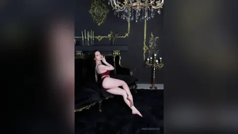 AniParadiser poses on a black chaise lounge wearing a red bikini, showing her long legs and dark hair in a dark, gold-accented room.