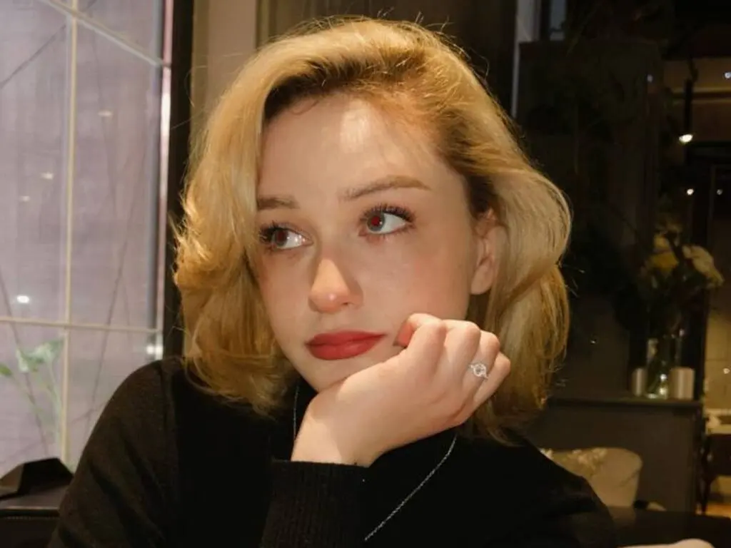 Blonde woman in a black turtleneck sweater looking thoughtful in a dimly lit indoor setting.