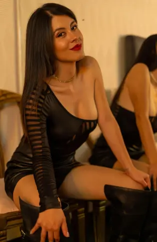 Glamorous woman in a black long-sleeve cutout dress sitting in a warm, moody indoor setting.