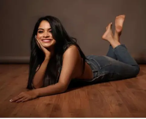 Smiling young woman in denim jeans lying on a wooden floor in a cozy, warm indoor setting.