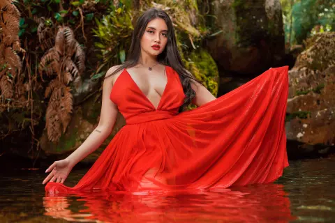 Elegant woman in a flowing red gown sitting in a lush jungle pond surrounded by tropical greenery.