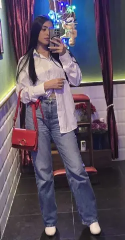 Young woman in white button-down shirt and blue jeans taking a mirror selfie in a cozy room.