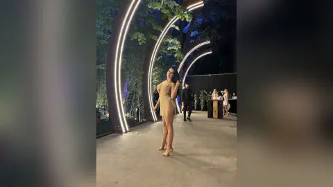 NicoleBella stands outdoors in a short tan dress, posing next to glowing light arches.
