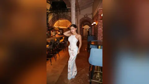 Elegant woman in a white halter top and floral trousers posing in a warm, rustic Mediterranean restaurant.