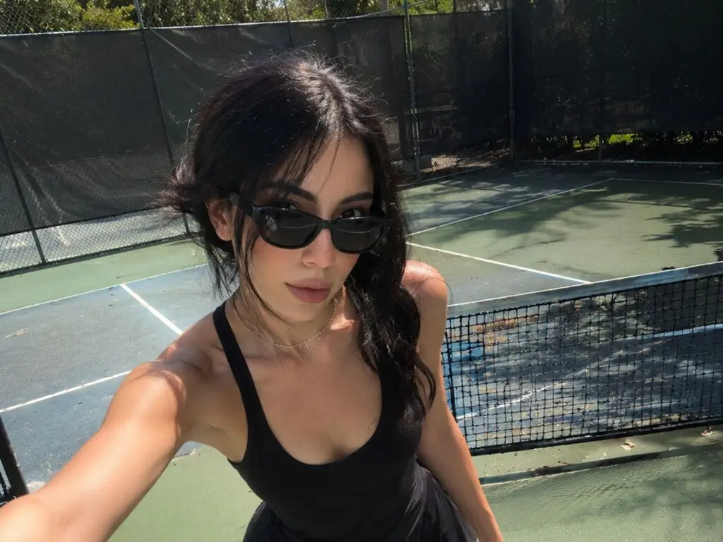 Young woman in black tank top and sunglasses relaxing on a sunny outdoor tennis court.