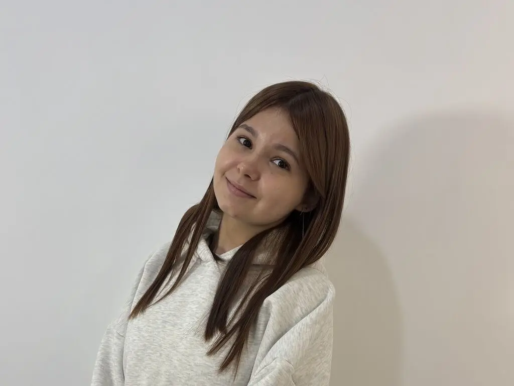 ChantelGloss tilts her head and smiles while wearing a light grey hooded sweatshirt against a plain white wall.