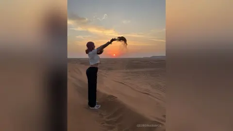 Woman in white crop top and black pants throwing sand in a vast desert landscape during a golden sunset.