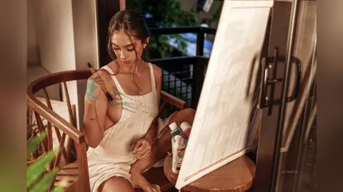 LucyMartin sits in a wooden chair wearing a white tank top while holding a paintbrush near her bare shoulder.