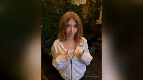 Young woman in a white Diesel zip-up hoodie smiling in a cozy outdoor garden setting.