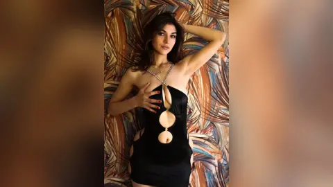 Elegant woman in a black cutout cocktail dress posing on a patterned fabric background with a glamorous mood.