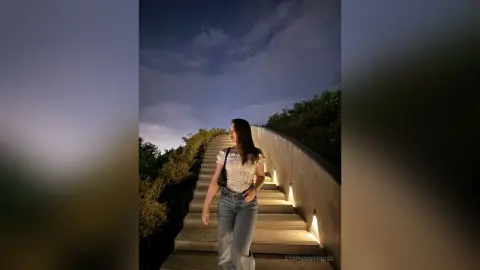 StephanieRiggi stands on outdoor stairs at night wearing a white top and blue jeans under a cloudy sky.