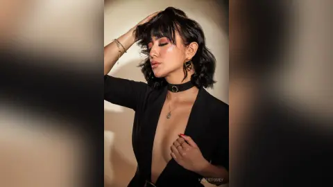 Elegant woman in a black plunging jumpsuit and gold jewelry posing with a sophisticated mood against a neutral wall.