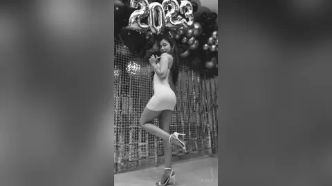 JessieLiu stands in profile wearing a white mini dress and high heels against a backdrop of silver sequins and black balloons.