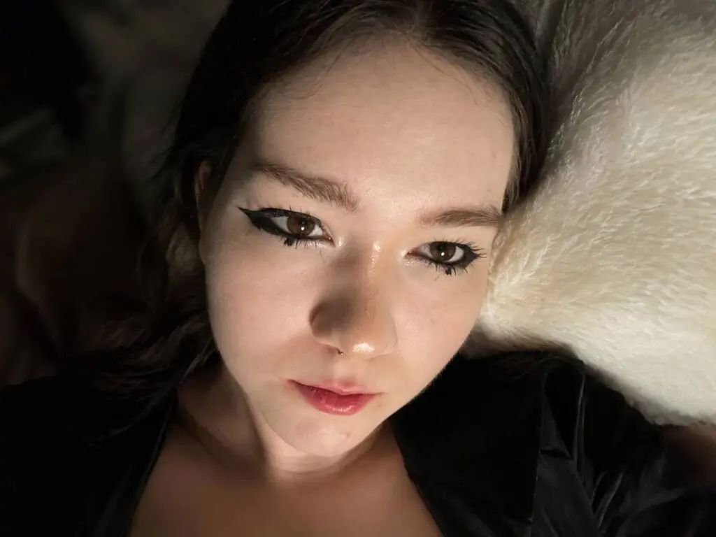 KendalChalet lying on a white pillow with her dark hair, wearing a black top and heavy black eyeliner.