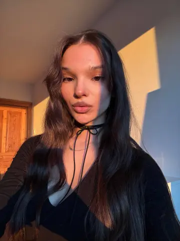 Noirbeauty poses with long wavy hair wearing a black v-neck top and a thin black choker in warm sunlight.