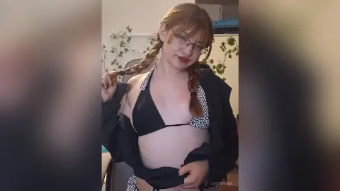 LucyMoon wears a black bikini top and glasses while posing with her braided hair in a room with ivy plants.