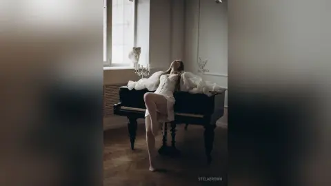 StelaBrown reclines back against a black piano wearing a white lace corset and matching white lace undies.