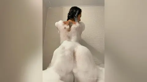 AngieSummers — Woman with back tattoo covered in white soap bubbles in a bright bathroom setting.