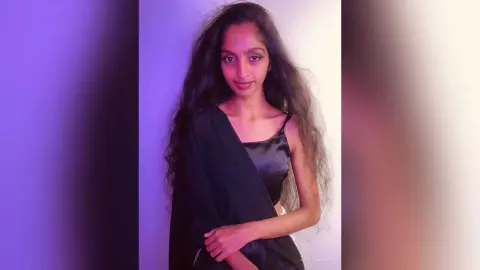 Smiling young woman with long wavy hair wearing a black satin traditional outfit against a plain background.