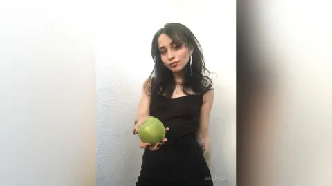 Young woman in a black tank top holding a green apple against a white wall in a moody, minimalist setting.