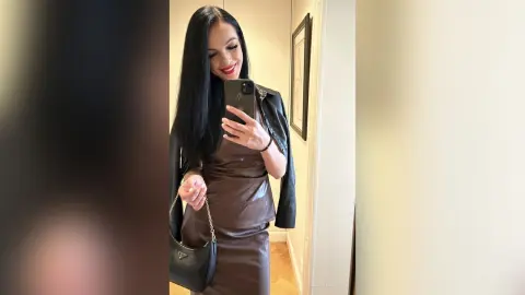 Smiling woman in a brown satin slip dress and black leather jacket holding a black purse in a bright hallway.