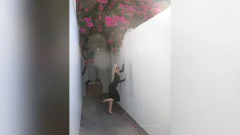 EllieFrosty stands against a white wall in a black dress, posing with one hand up near her blonde hair.