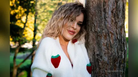 Blonde woman in a white strawberry knit sweater leaning against a tree in a sunny outdoor garden setting.