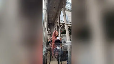 AmelieLautrec with long red hair sits at a glass table under a stone archway, holding a white cup and looking up.