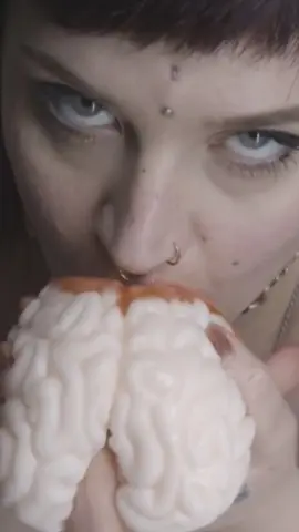 Close-up of a woman with bangs eating a brain-shaped dessert in a moody, artistic setting.