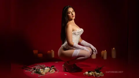 Elegant woman in white lace corset and gloves sitting against a red backdrop with glowing candles.