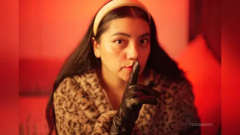 Young woman in leopard print coat and black gloves making a hush gesture in warm red lighting.