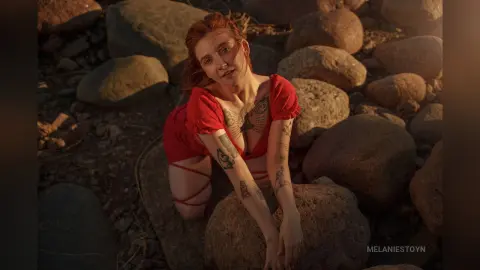 Redheaded woman in a red dress sitting among large rocks in warm, cinematic sunlight.