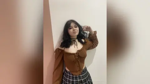 Young woman in a brown puff-sleeve top and plaid skirt posing against a neutral wall with a moody aesthetic.
