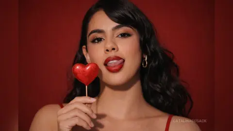 Young woman in a red outfit holding a glittery heart lollipop against a deep red background with a playful mood.