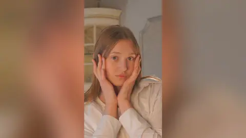 Blonde woman in a white shirt resting her face in her hands with a dreamy, thoughtful expression indoors.