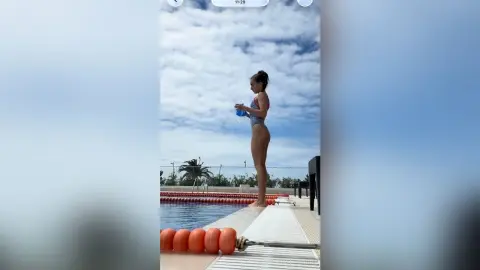 Fit woman in a purple swimsuit standing by a swimming pool under a bright blue sky.