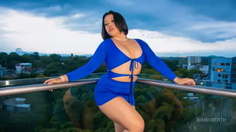 KimRoberth poses outdoors in a blue long-sleeved cutout bodysuit against a twilight sky and city view.
