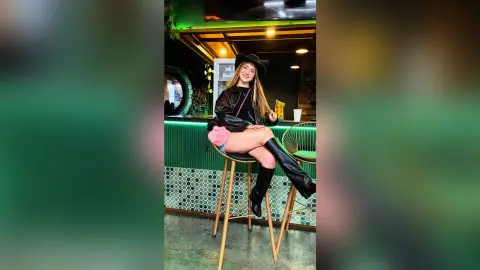 Stylish woman in black leather jacket and cowboy hat sitting on a bar stool in a modern green lounge.