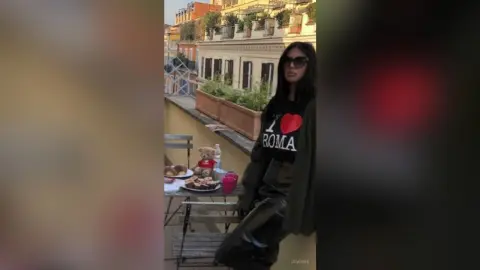 Stylish woman in a black graphic t-shirt relaxing on a sunny urban balcony with snacks and a teddy bear.