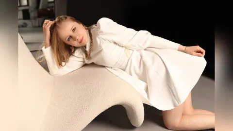 Elegant woman in a white long-sleeve dress leaning on a textured white sofa in a minimalist studio setting.