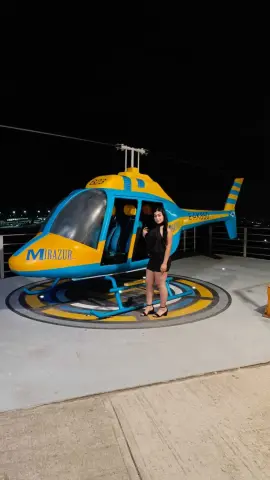 Stylish woman in a black dress standing next to a blue and yellow Mirazur helicopter at night.