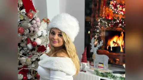 Smiling blonde woman in a white fuzzy holiday outfit posing near a decorated Christmas tree and fireplace.