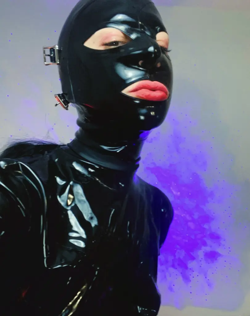 MissCocoDeville wears a black shiny mask and black straps, looking directly at the camera against a red background.