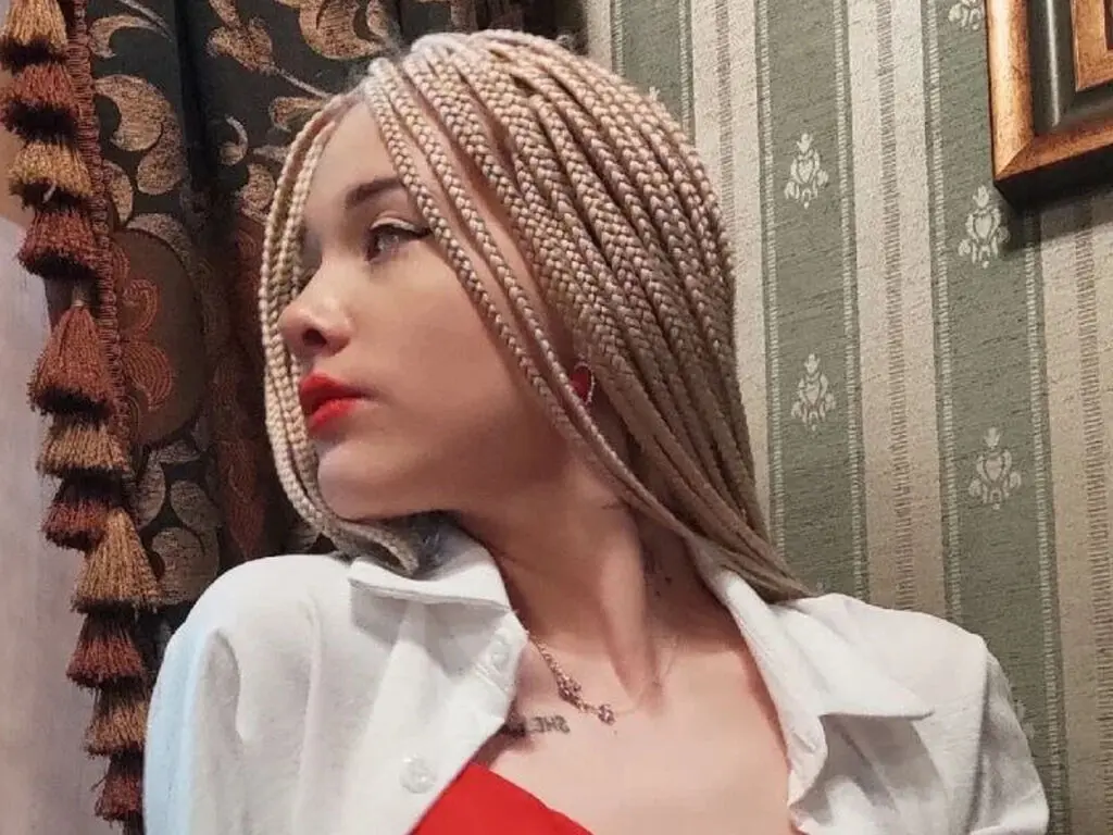 Young woman with blonde box braids and red lipstick wearing a white shirt in a vintage room.
