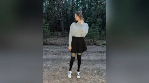 Young woman in a white sweater and black mini skirt posing in a moody, misty forest setting.
