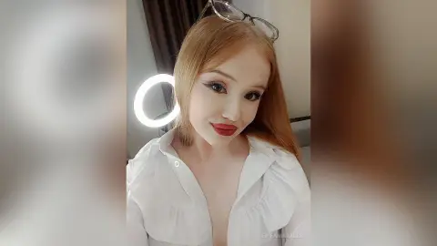 Blonde woman in a white ruffled blouse posing under ring light for a glamorous indoor portrait.