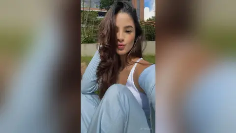 TefyRusso pouts her lips while wearing a white tank top and light blue sweater, showing off her long wavy brown hair outdoors.