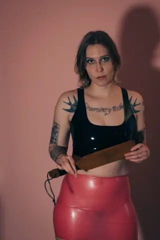 xoxoaliaquinn wears a black latex top and pink skirt while holding a wooden paddle against a pink background.