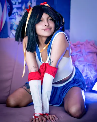 SakuraTao poses on a couch wearing a blue and white schoolgirl outfit with red armbands and black hair with yellow ribbons.