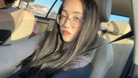 MayaOllin wearing glasses and a long-sleeved dark top, sitting in a car seat with her long black hair down.